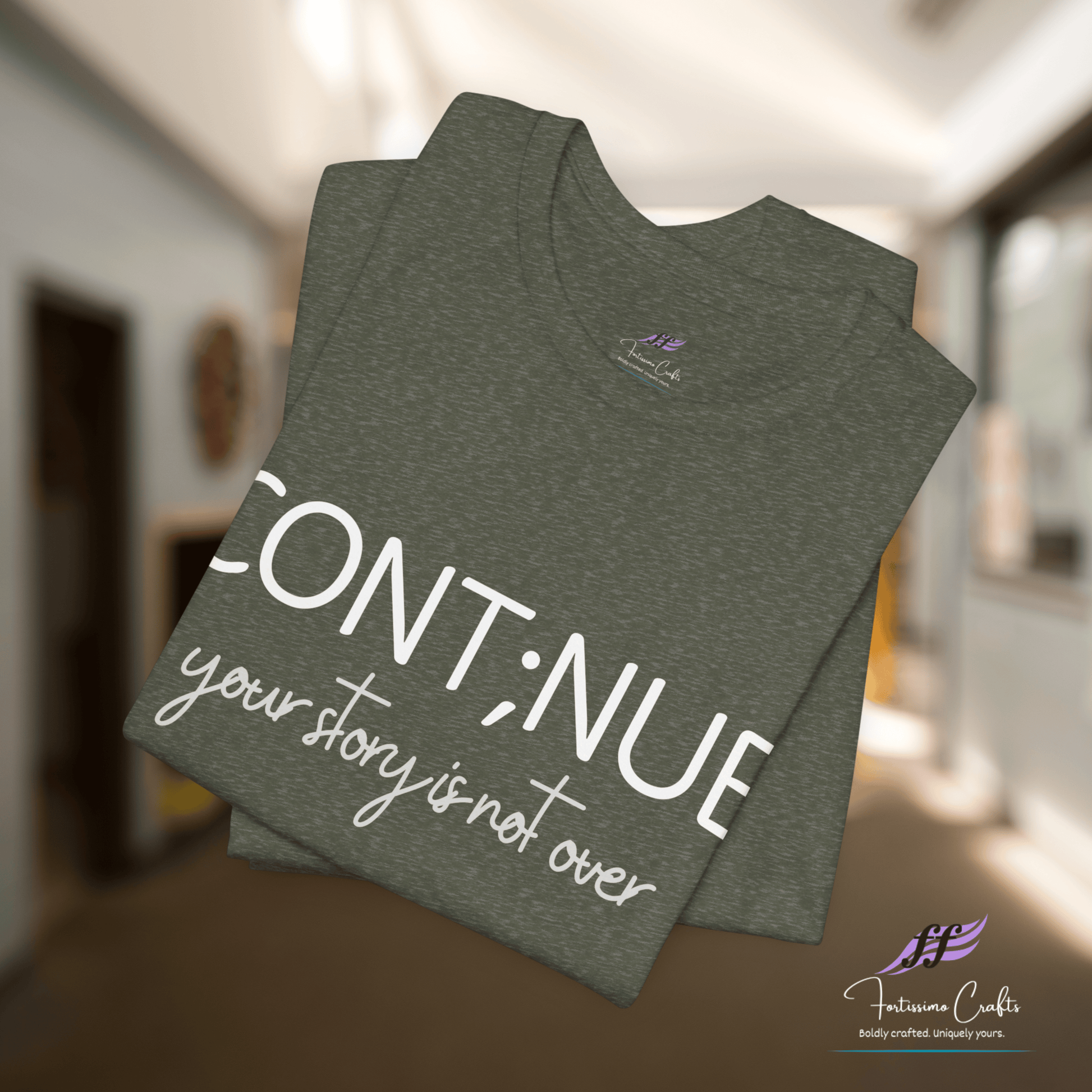 Folded dark gray t-shirt with 'CONT;NUE your story is not over' text in a blurred indoor setting.