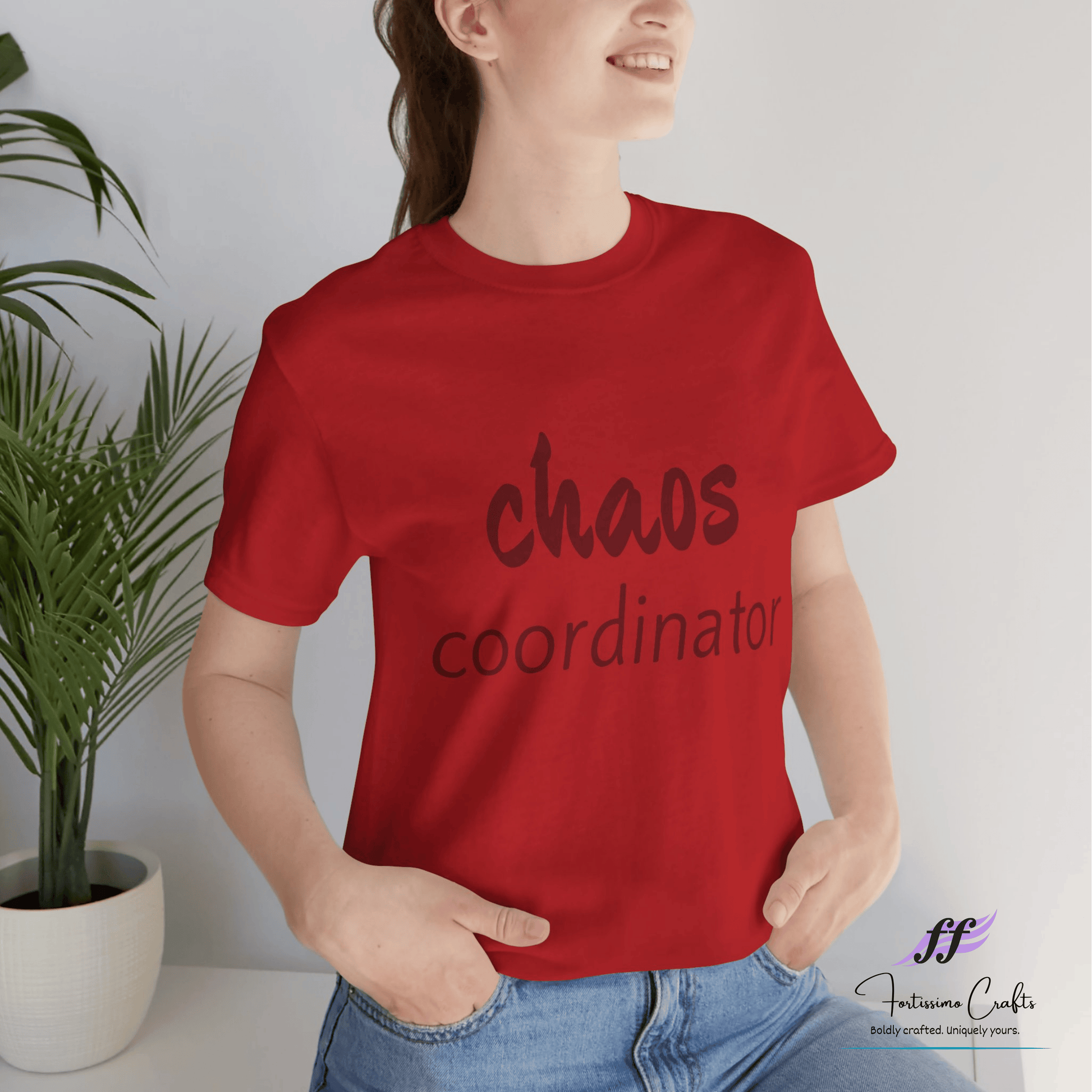 Person wearing a red t-shirt with 'chaos coordinator' text, standing against a white wall with a plant.