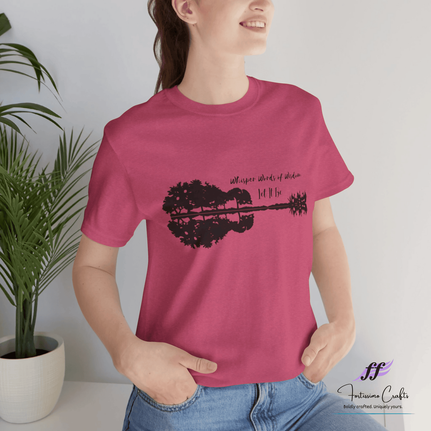 Person wearing a pink t-shirt with a black graphic design, standing indoors next to a plant.
