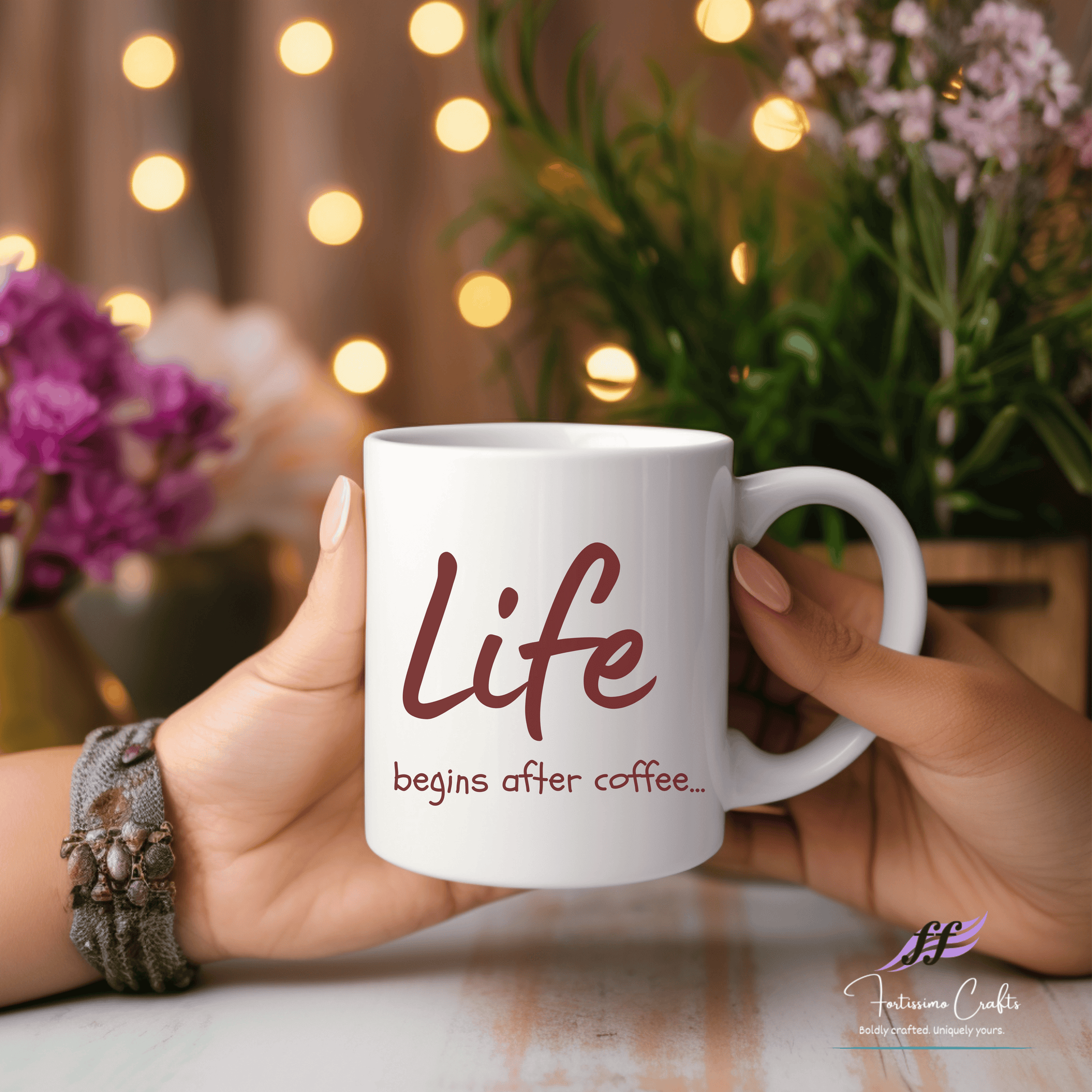 white ceramic mug with "Life begins after coffee..." in brown letters on the front held in both hands with flowers in the background